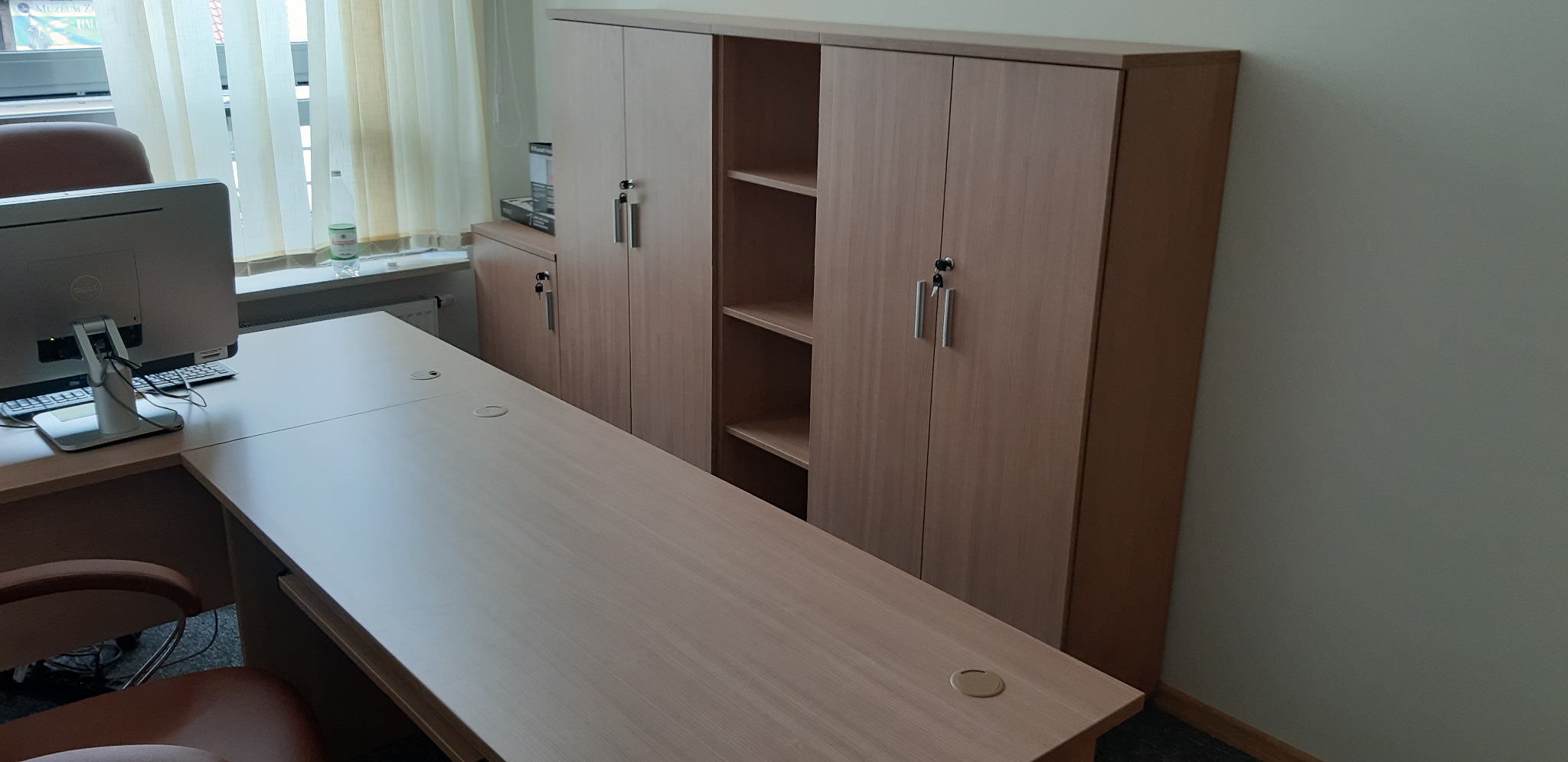 Jasne biuro z biurkiem narożnym i szafą zamykaną na klucz, widoczne gniazda w blacie biurka i komputer stacjonarny.