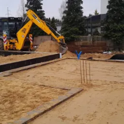 Żółta koparko-ładowarka JCB zasypuje piaskiem wykop pod fundamenty domu jednorodzinnego, widoczne zbrojenia i izolacja przeciwwilgociowa fundamentów.
