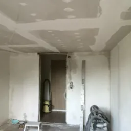 Wnętrze w trakcie prac wykończeniowych: sufit z płyt kartonowo-gipsowych z widocznymi łączeniami do szpachlowania, poziomica oparta o ścianę, szlifierka do gipsu oraz odkurzacz przemysłowy w rogu...
