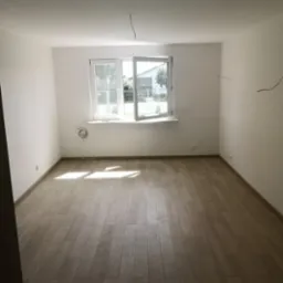 Pusty pokój po odświeżeniu z jasnymi panelami podłogowymi, białymi ścianami i otwartym oknem w słoneczny dzień. Widoczne wystające kable elektryczne.