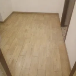 Ułożona podłoga z paneli imitujących drewno w pustym pokoju z widocznymi listwami przypodłogowymi i niedokończonym wykończeniem przy futrynie drzwi.