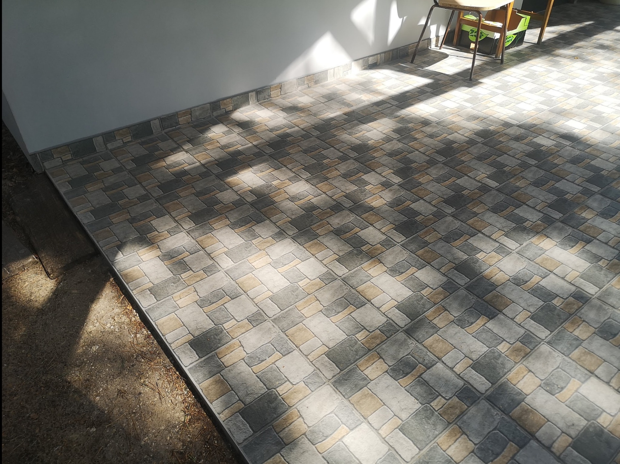 Ułożona podłoga z płytek ceramicznych w odcieniach szarości, beżu i brązu, z widocznym cokołem przy ścianie i fragmentem krzesła w tle, oświetlona promieniami słonecznymi tworzącymi kontrastujące...