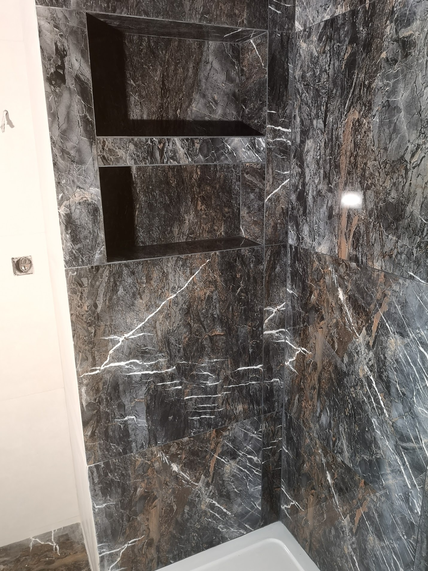 Ściana w łazience wyłożona ciemnymi płytkami imitującymi marmur z wnękami na kosmetyki, widoczny fragment białej ściany i wanny.