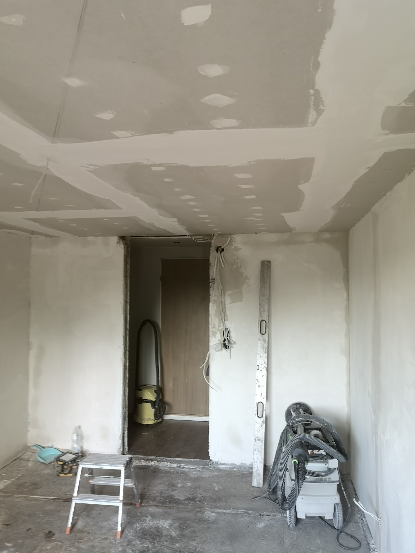 Wnętrze w trakcie prac wykończeniowych: sufit z płyt kartonowo-gipsowych z widocznymi łączeniami do szpachlowania, poziomica oparta o ścianę, szlifierka do gipsu oraz odkurzacz przemysłowy w rogu...