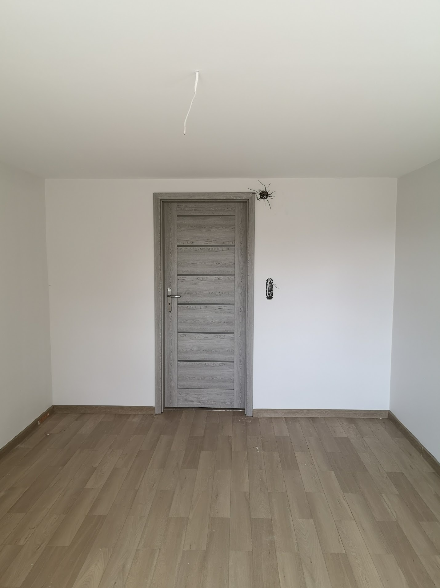 Pomalowane na biało wnętrze pokoju z panelami podłogowymi i szarą, drewnopodobną stolarką drzwiową; widoczne wystające kable i atrapa pająka na ścianie.