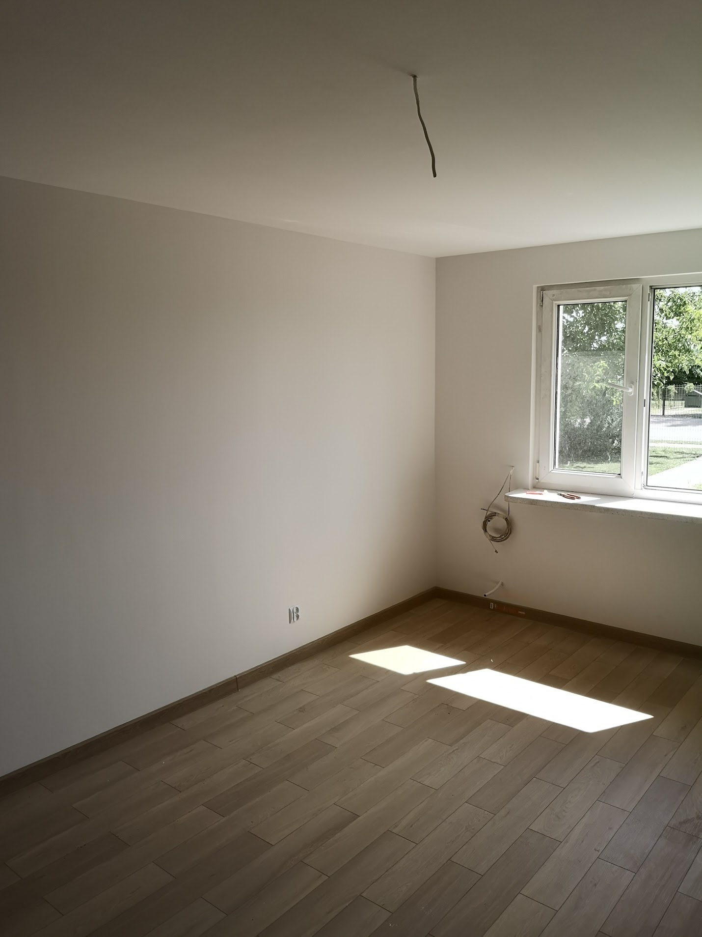 Pusty pokój z drewnopodobną podłogą, świeżo pomalowanymi ścianami w odcieniach bieli, oświetlony naturalnym światłem wpadającym przez okno, z widocznymi wystającymi kablami ze ściany i sufitu...