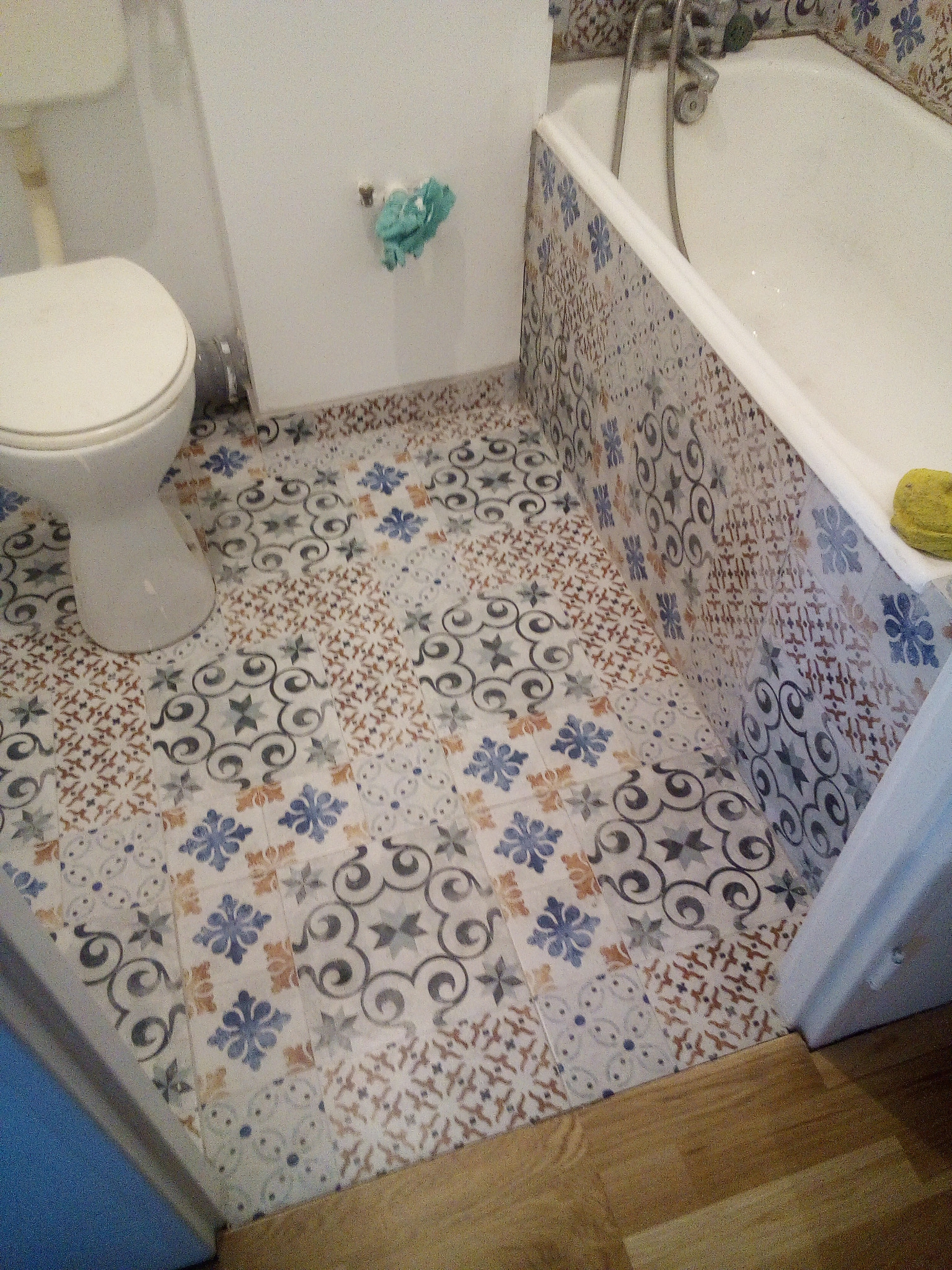 Biała toaleta i wanna obudowana płytkami z wzorem patchworkowym w łazience z podłogą wyłożoną panelami imitującymi drewno.