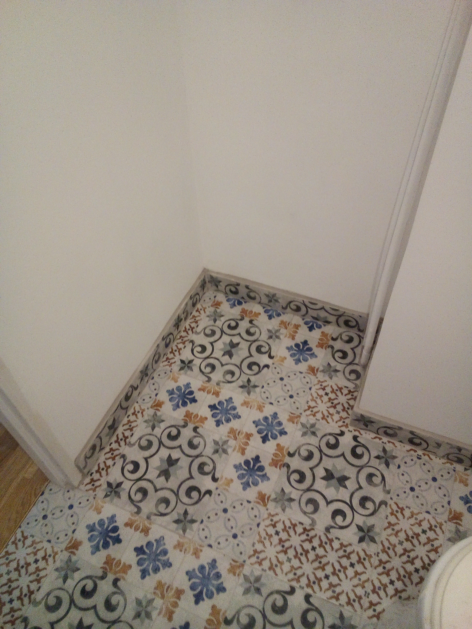 Wzorzysta podłoga z płytek ceramicznych w rogu łazienki, widok z góry, z fragmentem białej ściany i ościeżnicy.