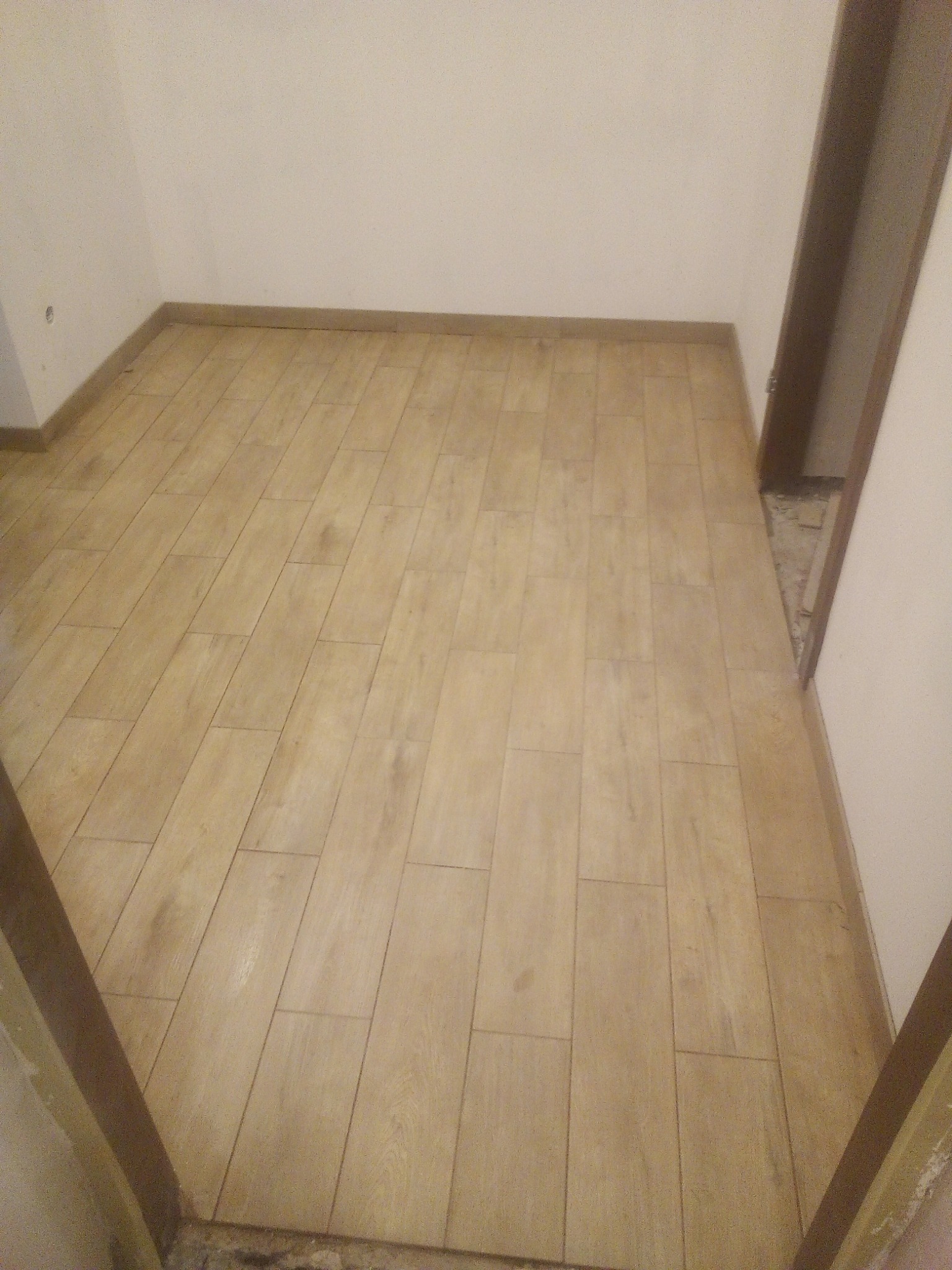 Ułożona podłoga z paneli imitujących drewno w pustym pokoju z widocznymi listwami przypodłogowymi i niedokończonym wykończeniem przy futrynie drzwi.