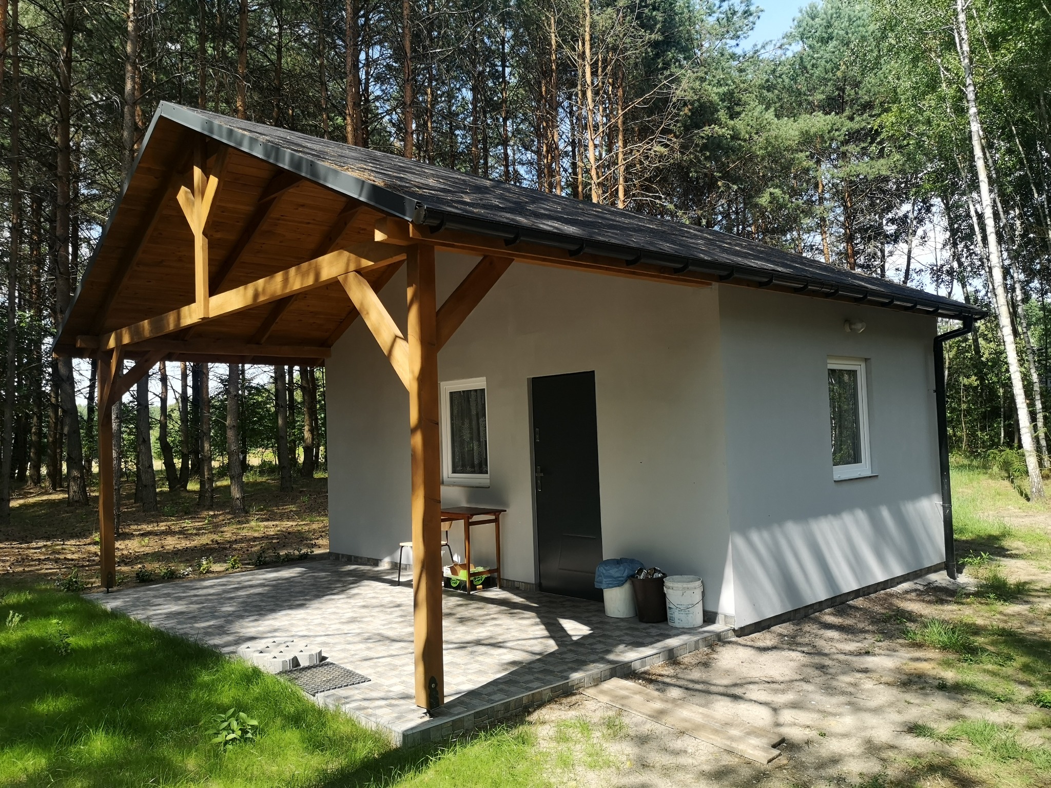 Nowo wybudowany, parterowy budynek z szarą elewacją i drewnianą konstrukcją zadaszenia tarasu, otoczony sosnowym lasem, z widocznymi wiaderkami po farbie i narzędziami na zewnątrz.