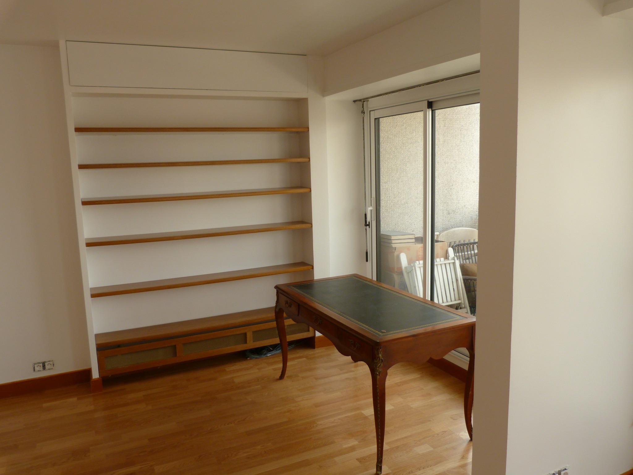 Pusty pokój z wbudowanym regałem z drewnianymi półkami, biurkiem z zielonym blatem i wyjściem na balkon.