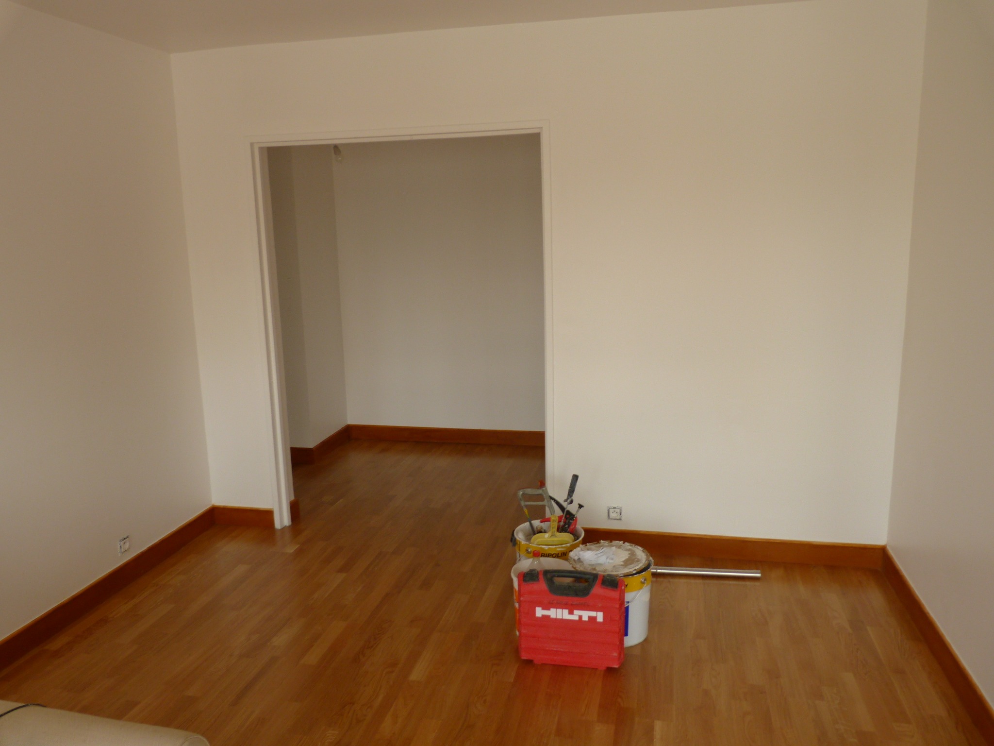 Wnętrze pokoju po malowaniu ścian na biało, widoczne narzędzia malarskie, kuweta z farbą oraz czerwona skrzynka narzędziowa Hilti na parkiecie przy ścianie.
