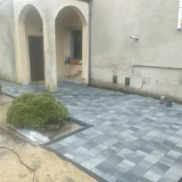 Ułożona szara kostka brukowa na posesji, widoczny fragment budynku z łukowatymi wejściami i niewielki krzew iglasty.