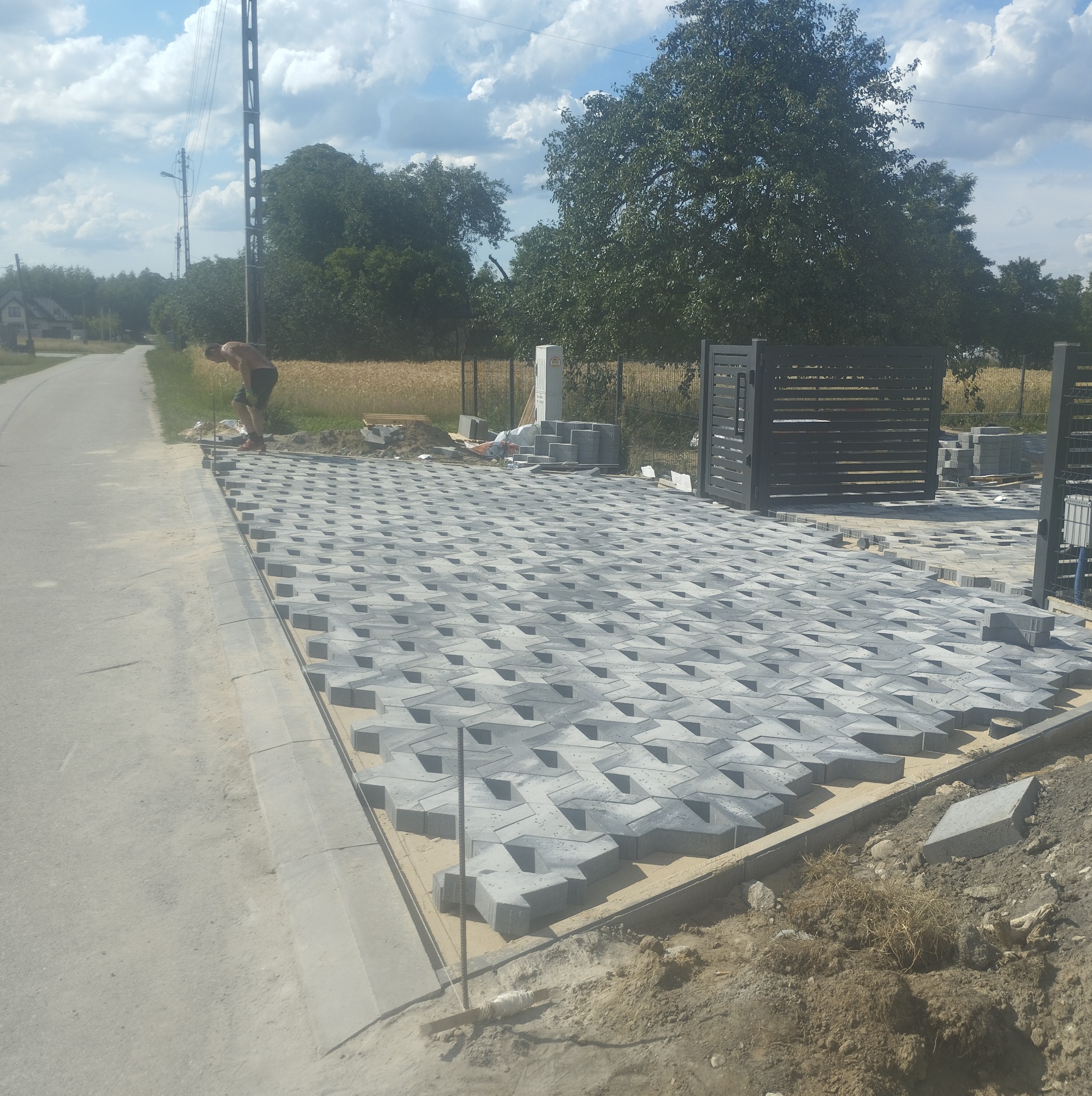 Układanie szarej kostki brukowej o specyficznym wzorze na posesji, widoczny pracownik przy krawędzi oraz przygotowane materiały budowlane w tle, częściowo ukończona nawierzchnia.