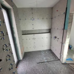 Wnętrze łazienki w trakcie kładzenia szarych płytek z użyciem krzyżyków dystansowych; widoczny fragment ściany z dekoracyjną, heksagonalną mozaiką oraz przymocowany odpływ liniowy.