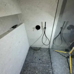 Szara łazienka z kabiną prysznicową typu walk-in, czarna armatura natryskowa, płytki podłogowe w odcieniach szarości, widoczny statyw poziomujący.