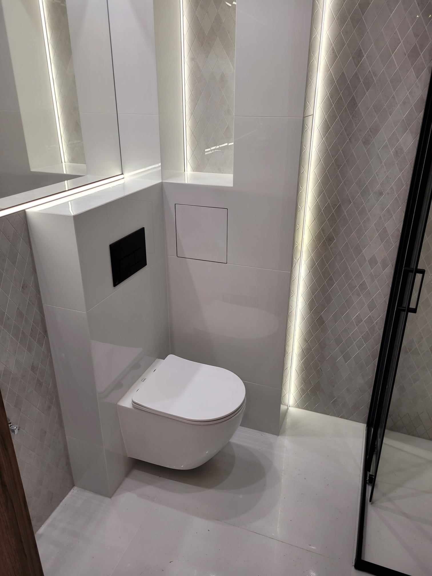 Nowoczesna łazienka z podwieszaną toaletą, płytkami w romby i oświetleniem LED wzdłuż ścian, widok na kabinę prysznicową z czarną ramą.
