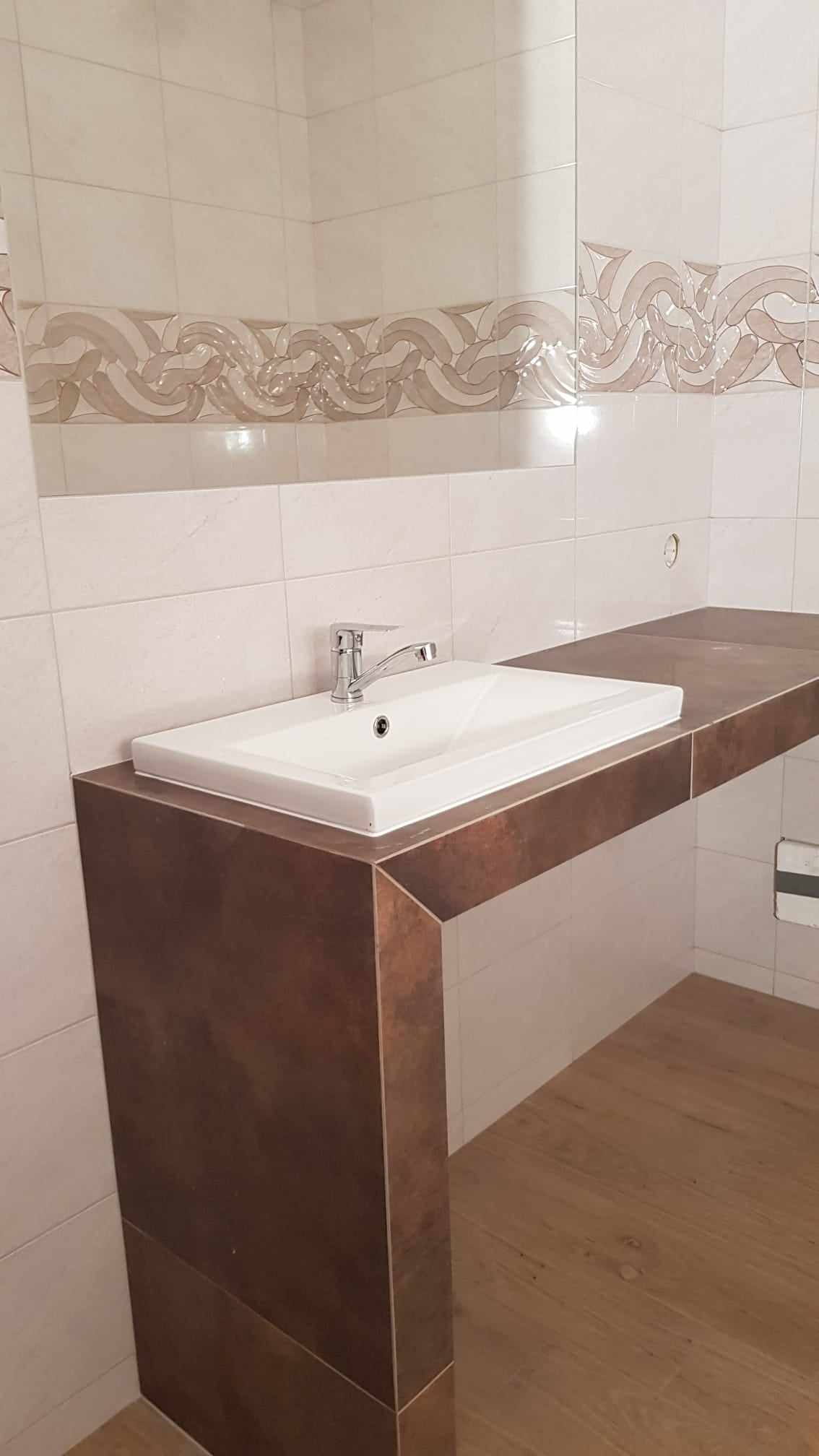 Nowoczesna łazienka z białą umywalką nablatową na brązowym blacie z płytek, kremowe płytki ścienne z dekoracyjnym pasem, drewniana podłoga.