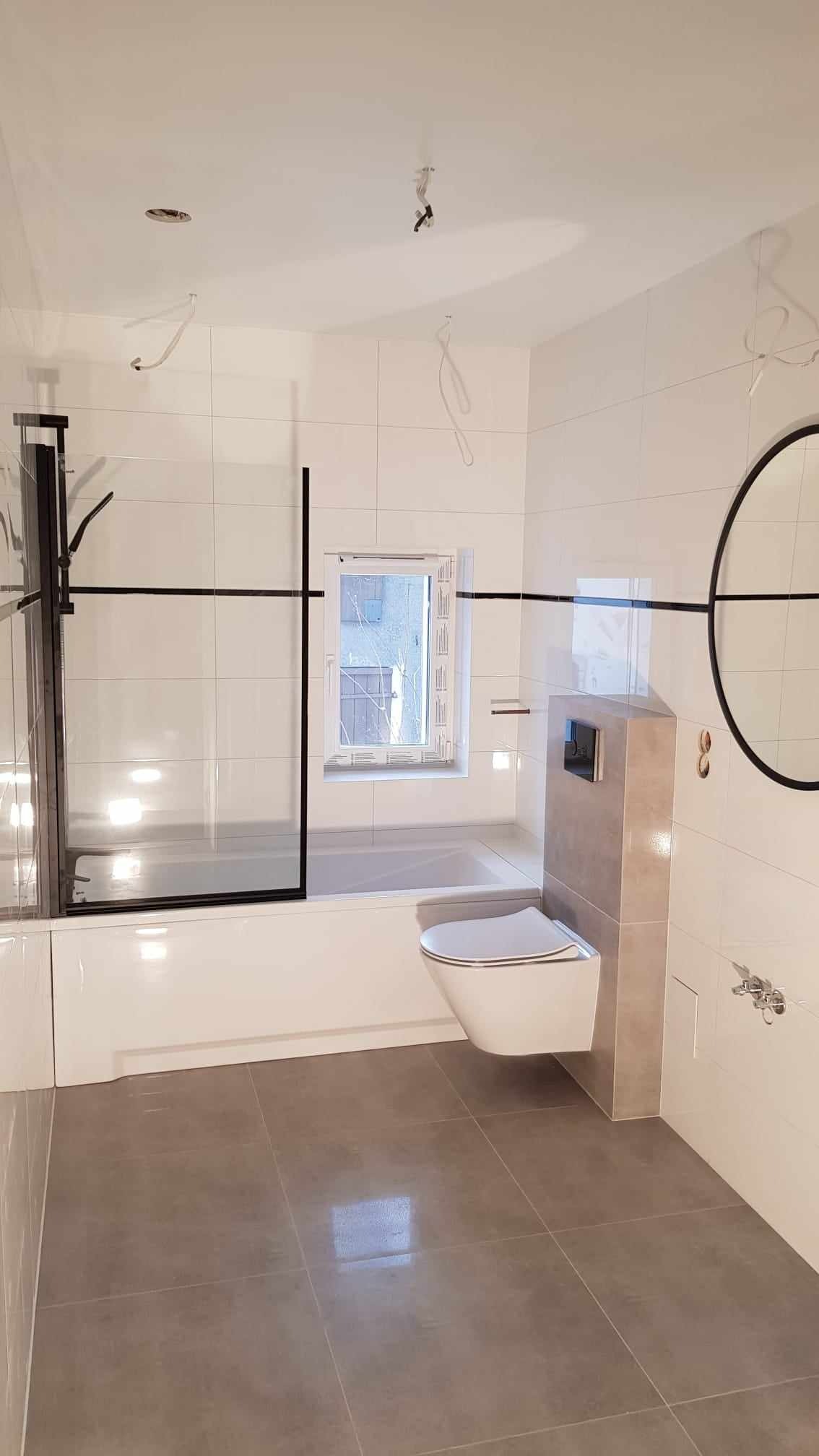 Nowoczesna łazienka z białymi płytkami ściennymi, szarą podłogą, wiszącą toaletą, kabiną prysznicową z czarnymi ramami i okrągłym lustrem. Widoczne niedokończone podłączenia elektryczne na suficie.