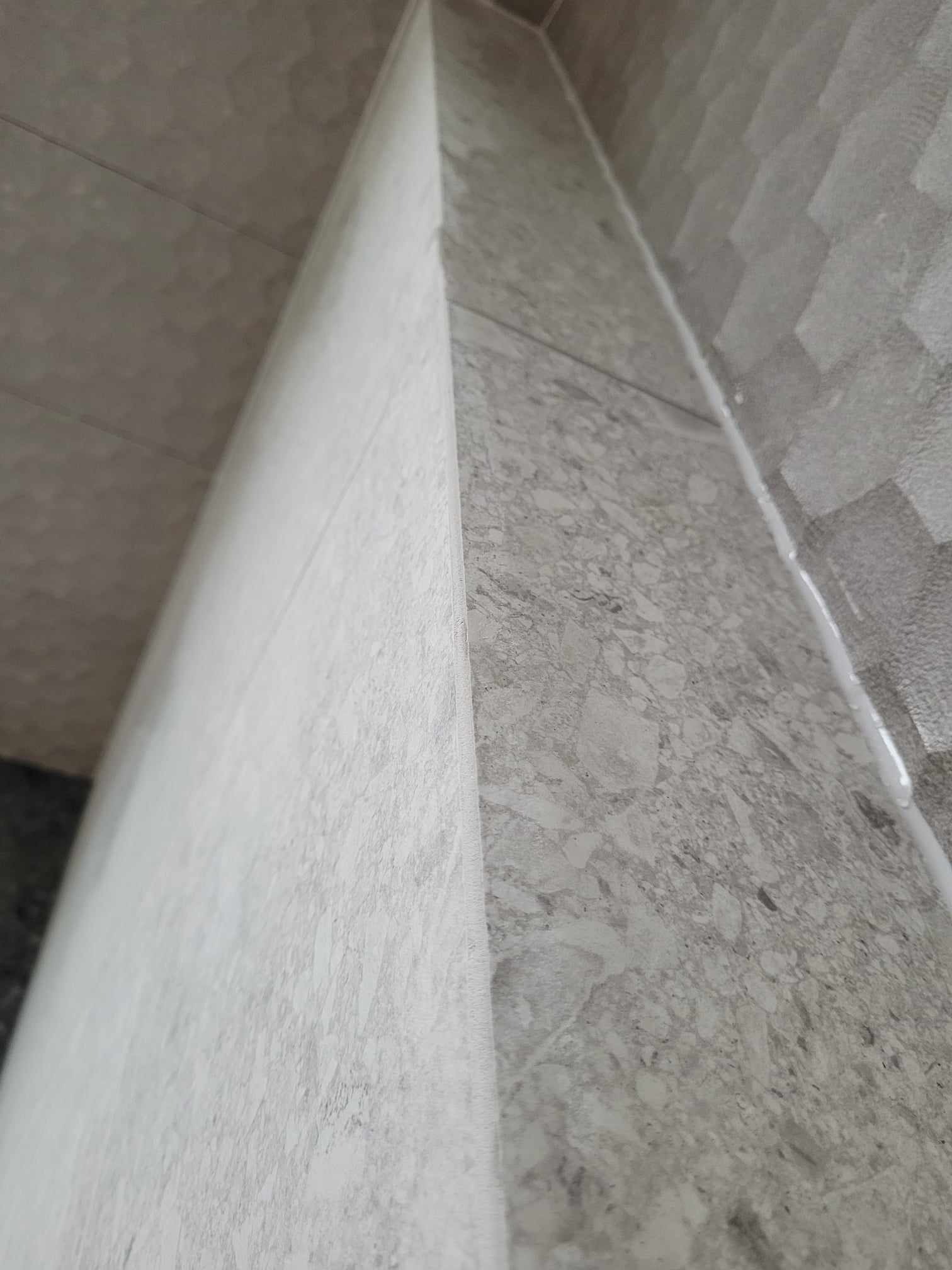 Świeżo położone, szare płytki imitujące marmur na ścianie, z widoczną fugą silikonową w rogu, sfotografowane pod kątem.