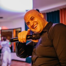 EL Wariat - Uśmiechnięty, łysy mężczyzna w czarnej koszuli śpiewa do mikrofonu na tle sali bankietowej z tańczącymi gośćmi. Dynamiczne ujęcie z bliska.