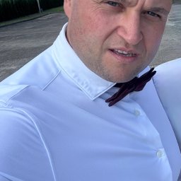 EL Wariat - Portret mężczyzny w białej koszuli i bordowej muszce na tle błękitnego nieba i zieleni. Ujęcie z dołu, podkreślające pewność siebie i profesjonalizm.