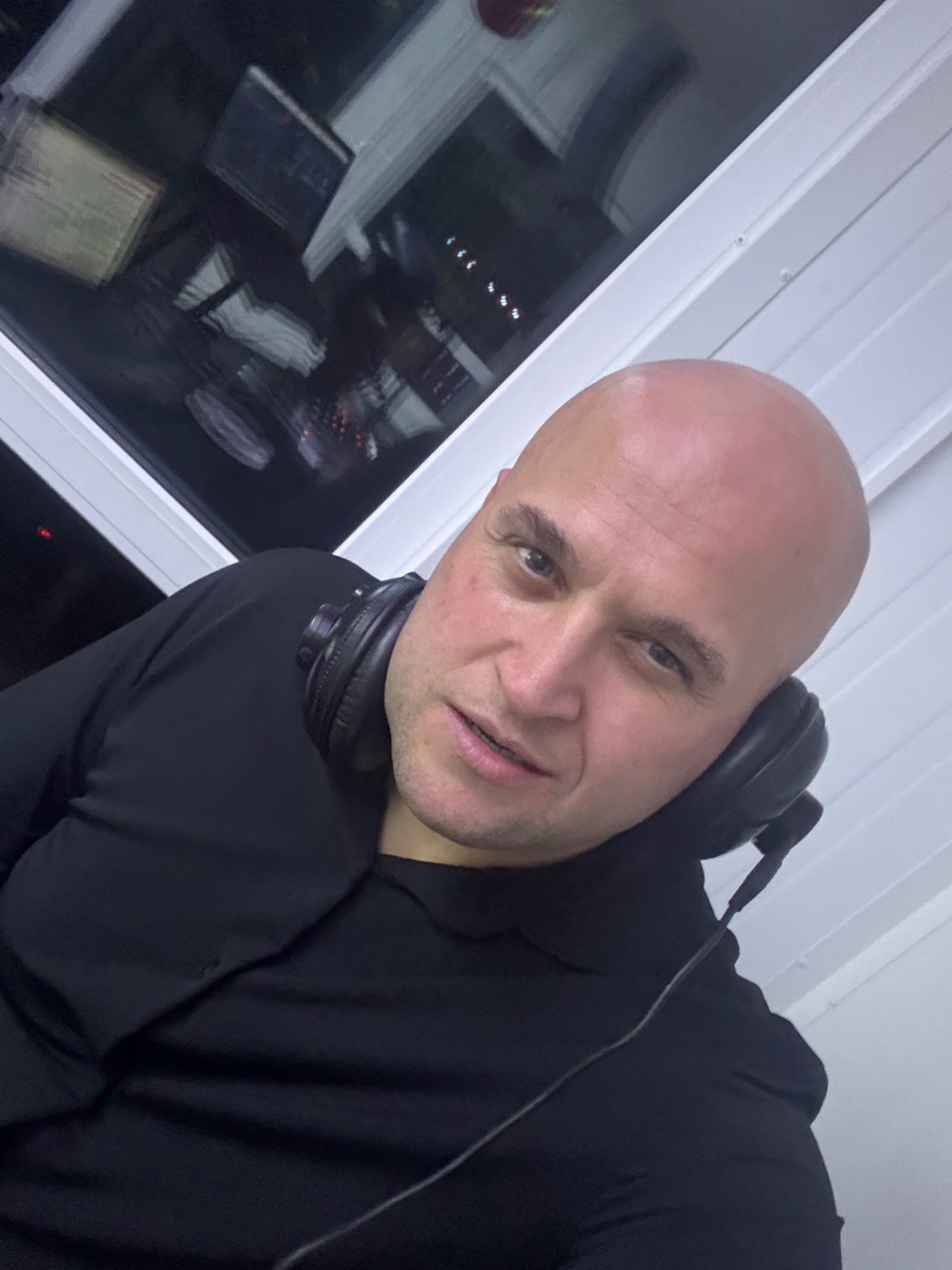 Portret mężczyzny z ogoloną głową i słuchawkami na szyi, w czarnej koszuli, z widocznym oknem w tle. Ujęcie z dołu.