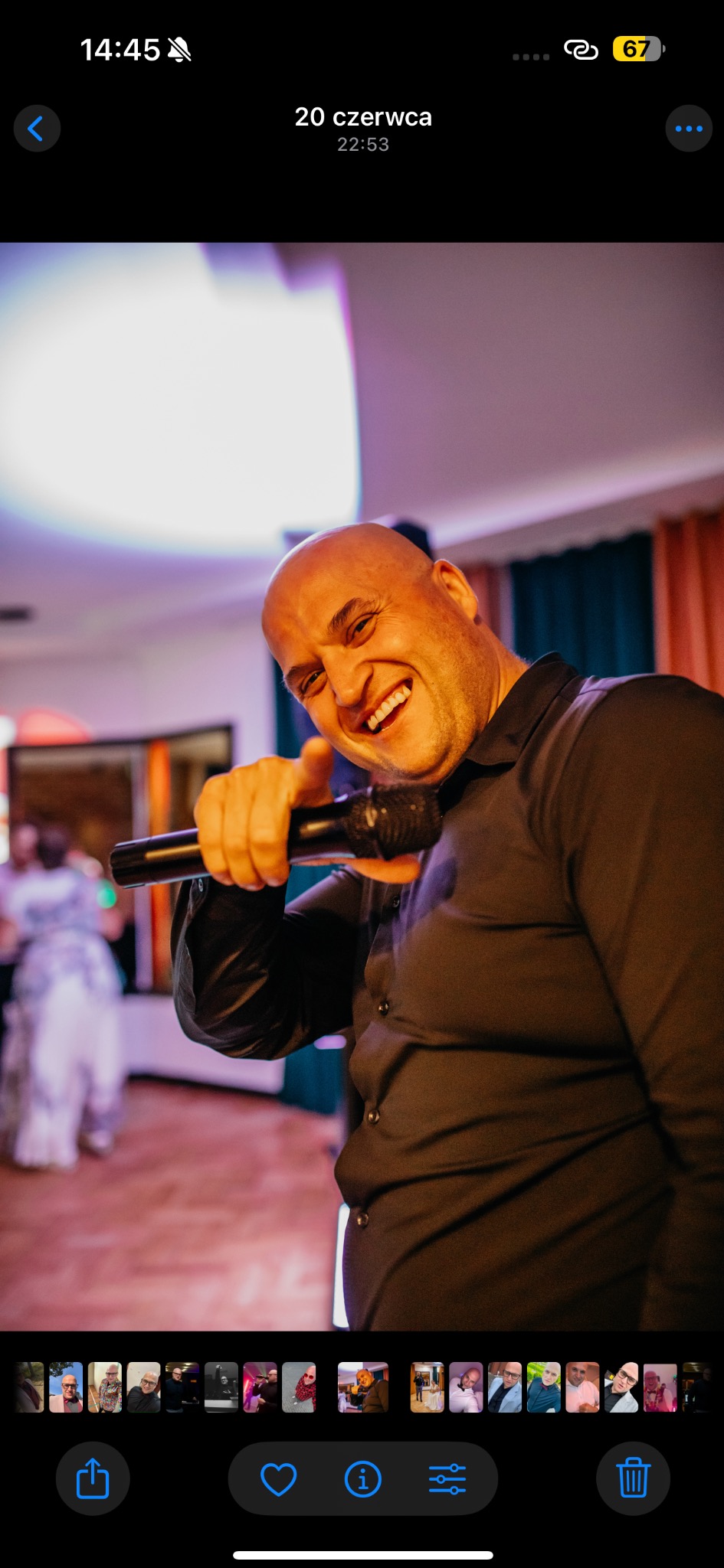 Uśmiechnięty, łysy mężczyzna w czarnej koszuli śpiewa do mikrofonu na tle sali bankietowej z tańczącymi gośćmi. Dynamiczne ujęcie z bliska.