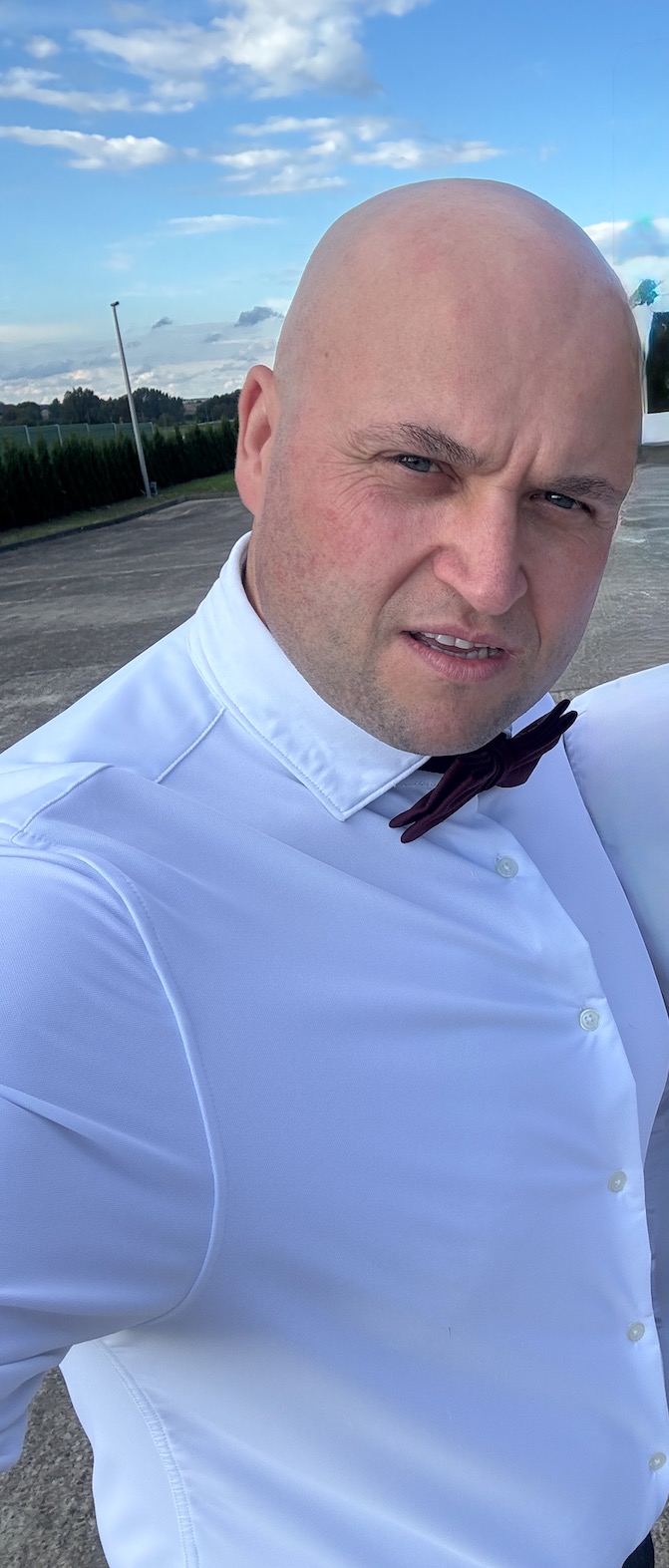 Portret mężczyzny w białej koszuli i bordowej muszce na tle błękitnego nieba i zieleni. Ujęcie z dołu, podkreślające pewność siebie i profesjonalizm.