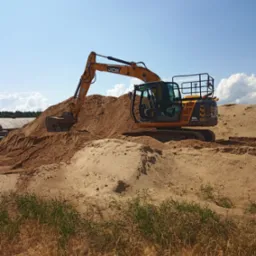 Żółta koparka JCB JS130LC na szczycie pryzmy piasku, z widocznymi śladami opon, na tle błękitnego nieba z białymi obłokami i zielenią w dolnej części kadru.