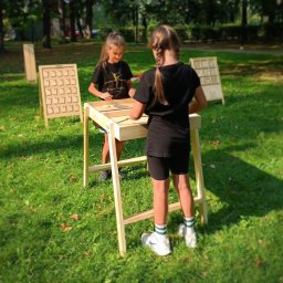 Nexturniej - atrakcje i animacje dla dzieci i dorosłych - Dwie dziewczynki grają w gry planszowe na świeżym powietrzu, drewniany stół i plansze do gier na trawie w parku.