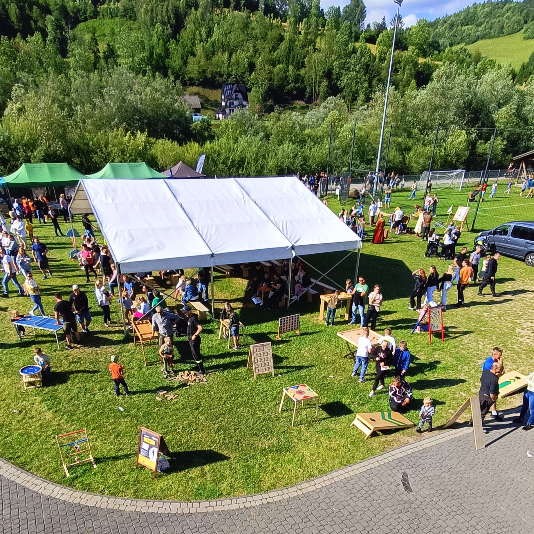 Zabawy plenerowe podczas imprezy na trawie: biały namiot, gry zręcznościowe, ping pong, uczestnicy w różnym wieku, tło leśne.