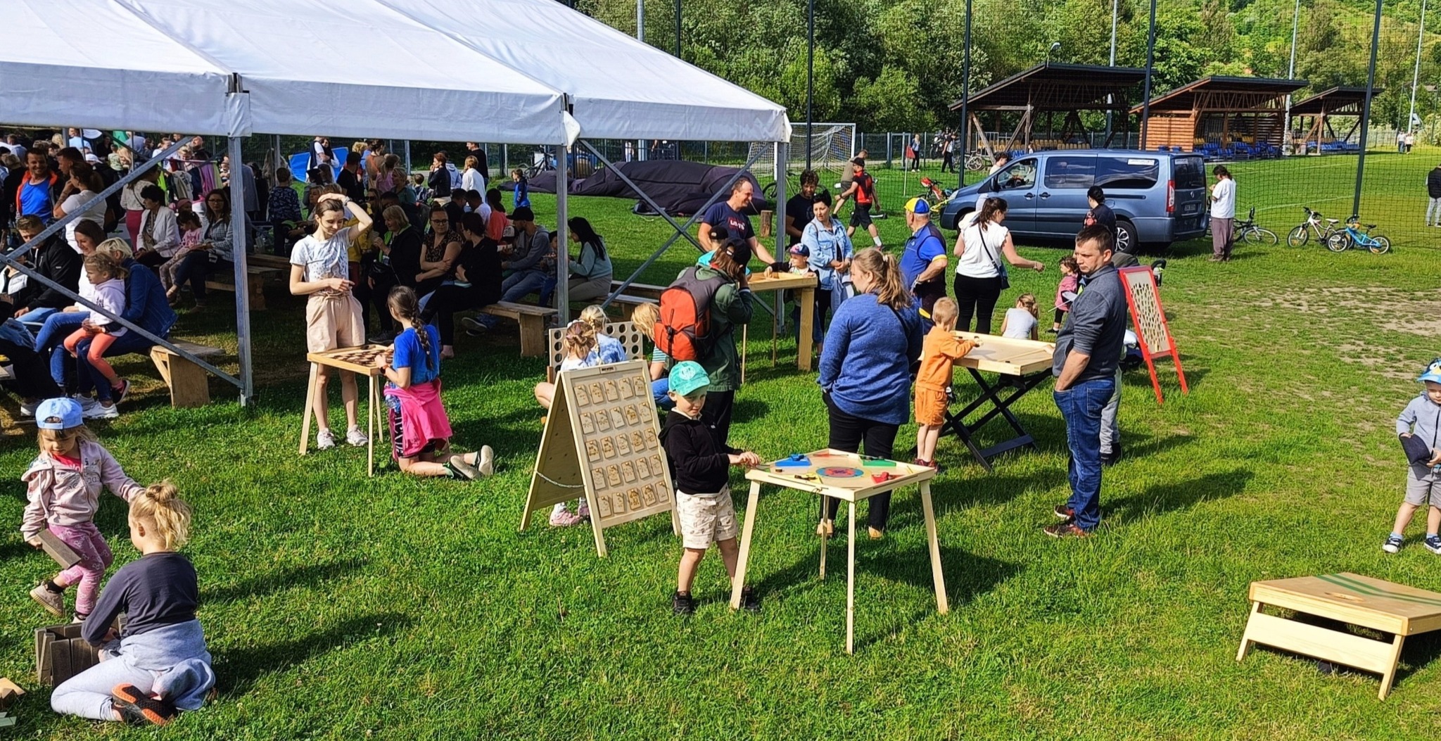Plenerowa impreza z drewnianymi grami edukacyjnymi dla dzieci, odbywająca się na trawie pod białym namiotem, z widocznymi grami zręcznościowymi i logicznymi oraz grupą dzieci i dorosłych.
