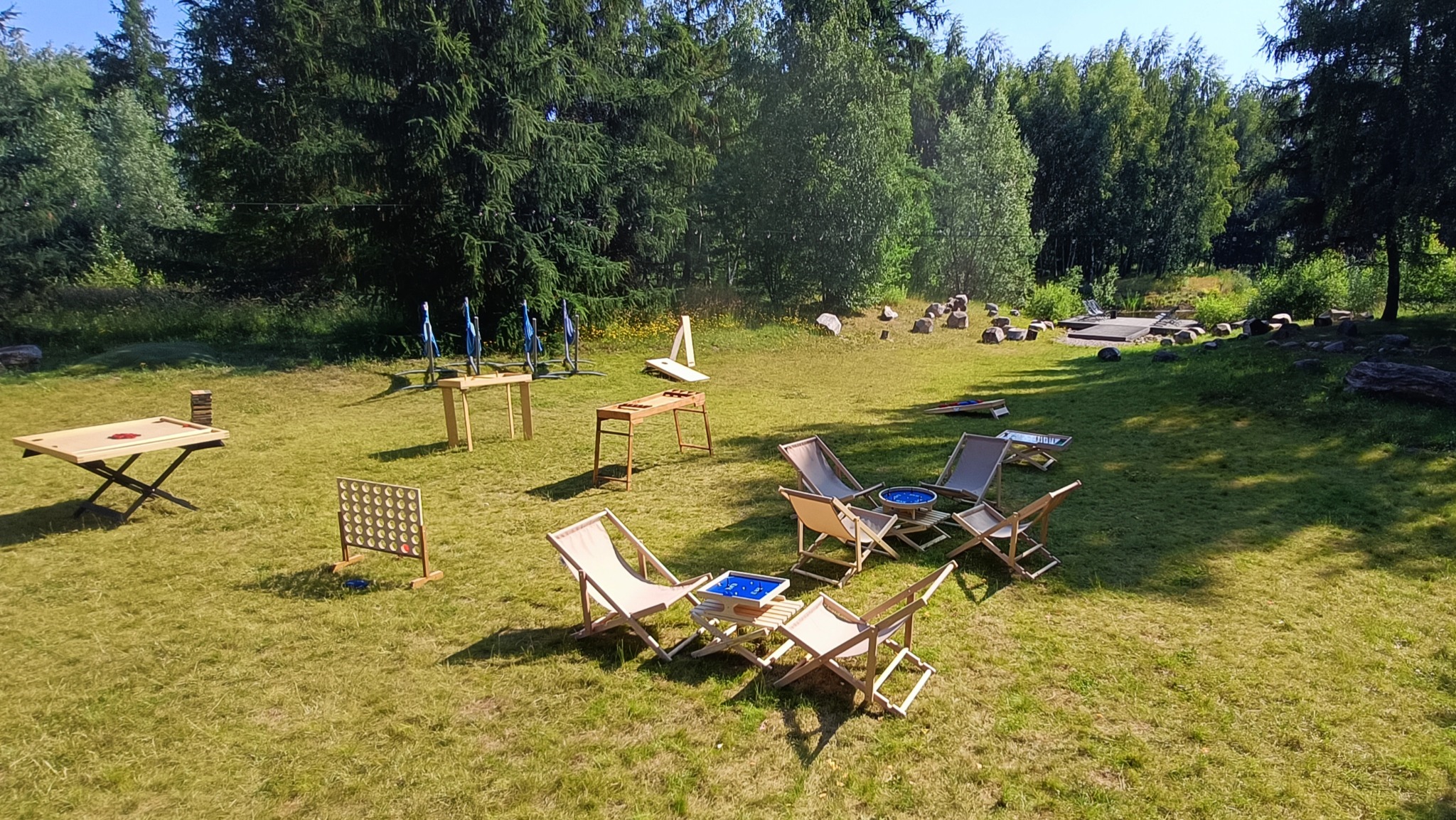 Plenerowa strefa relaksu z drewnianymi leżakami i grami: Jenga, Cztery w rzędzie, Cornhole, gra w rzutki, oraz stoliki. Tło stanowią drzewa i trawnik.