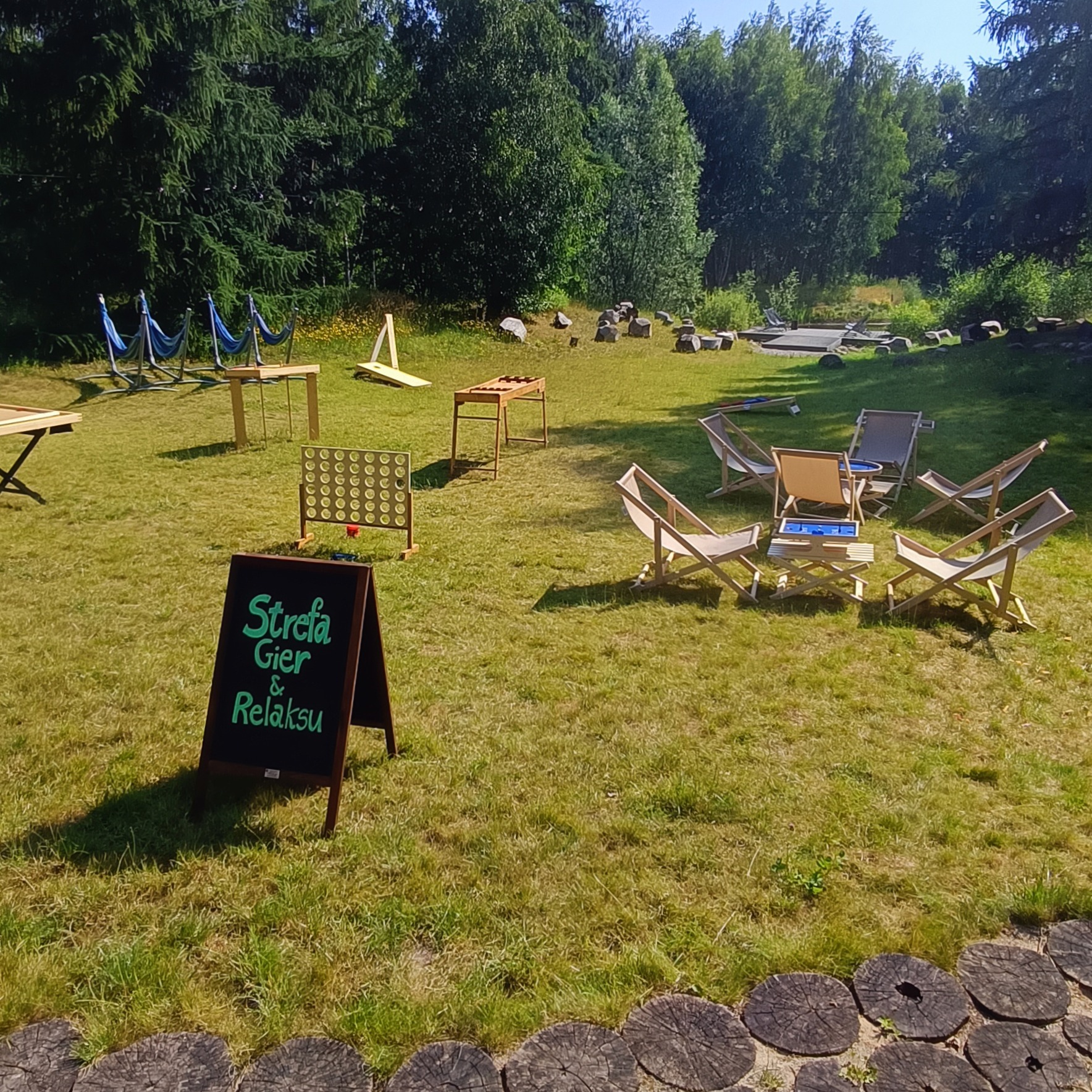 Zewnętrzna strefa gier i relaksu z hamakami, leżakami i drewnianymi grami na trawie, otoczona drzewami i oznaczona tablicą z napisem 'Strefa Gier & Relaksu'.