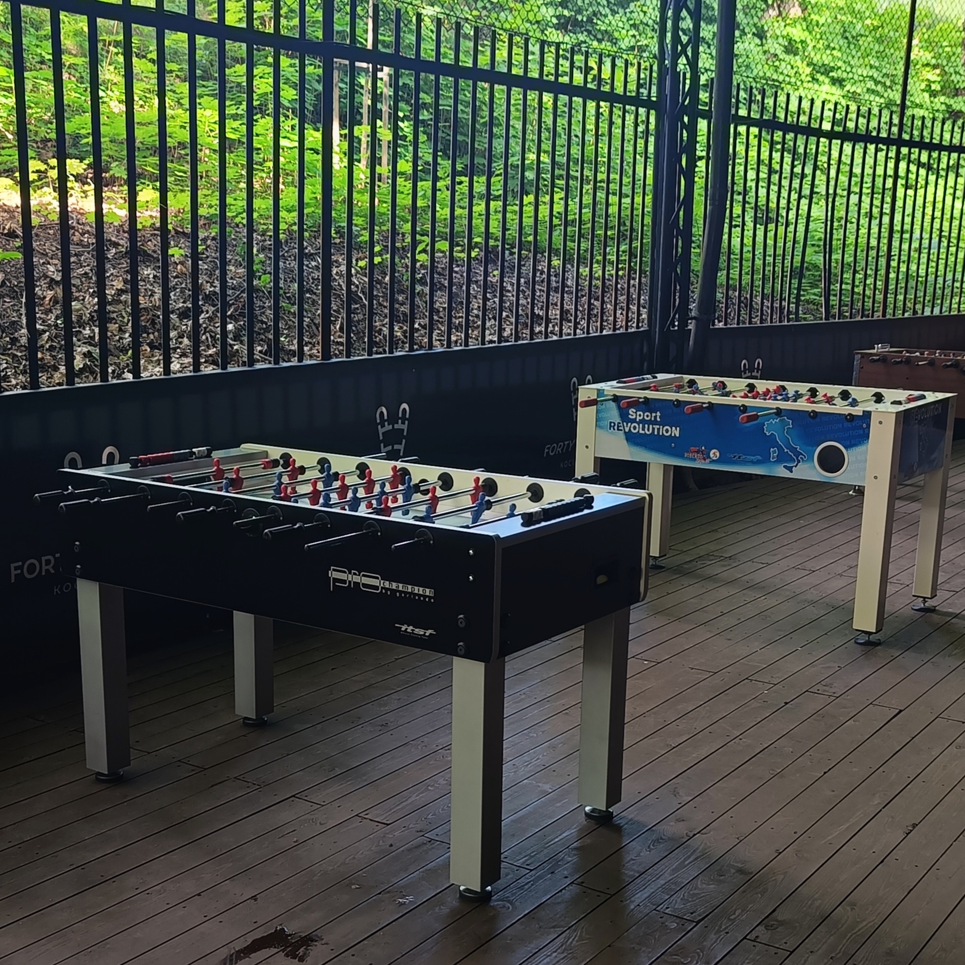 Dwa stoły do piłkarzyków na drewnianym tarasie z metalową barierką w tle, jeden czarny z napisem 'PRO champion by gariando', drugi biały z grafiką 'Sport Revolution'.