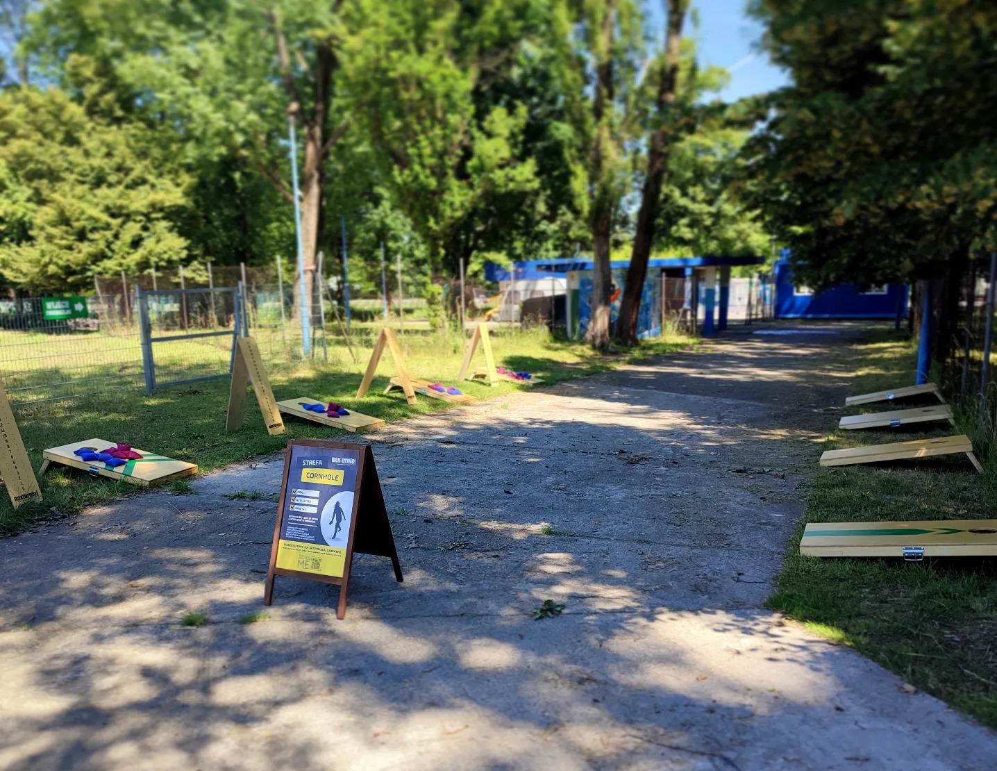 Zestawy do gry w cornhole ustawione na trawie w parku, z widoczną tablicą informacyjną 'Strefa Age Smile Cornhole', w tle drzewa i budynek.