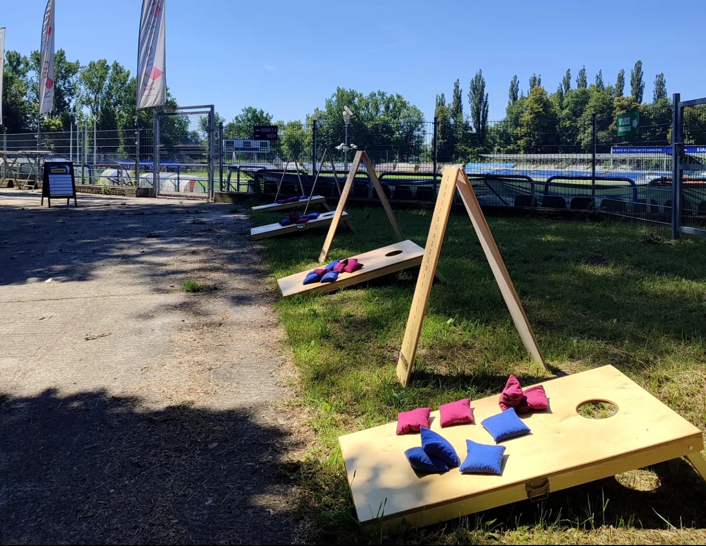 Dwie drewniane platformy do gry w rzut woreczkami (bean bag toss) ustawione na trawie, z niebieskimi i różowymi woreczkami, w tle ogrodzenie i drzewa.