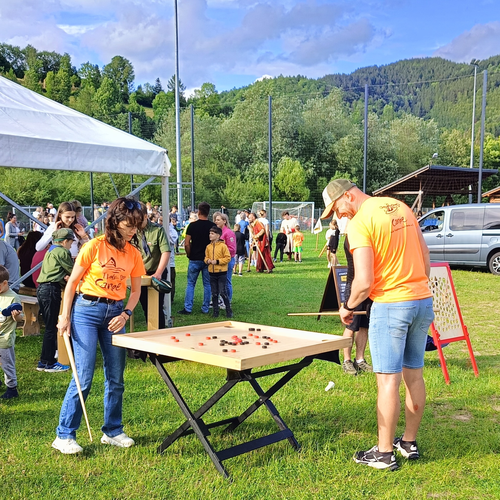 Uczestnicy w pomarańczowych koszulkach grają w grę z drewnianymi krążkami na stole podczas plenerowego wydarzenia z widocznymi górami w tle.
