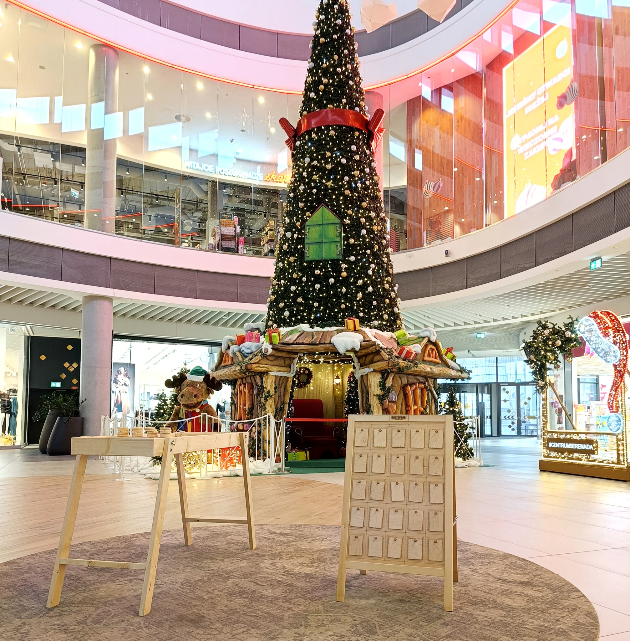 Dekoracja świąteczna w centrum handlowym: wysoka choinka z ozdobami, drewniany domek z oknem, pluszowy łoś w czapce Mikołaja oraz drewniane stoisko z kartkami.