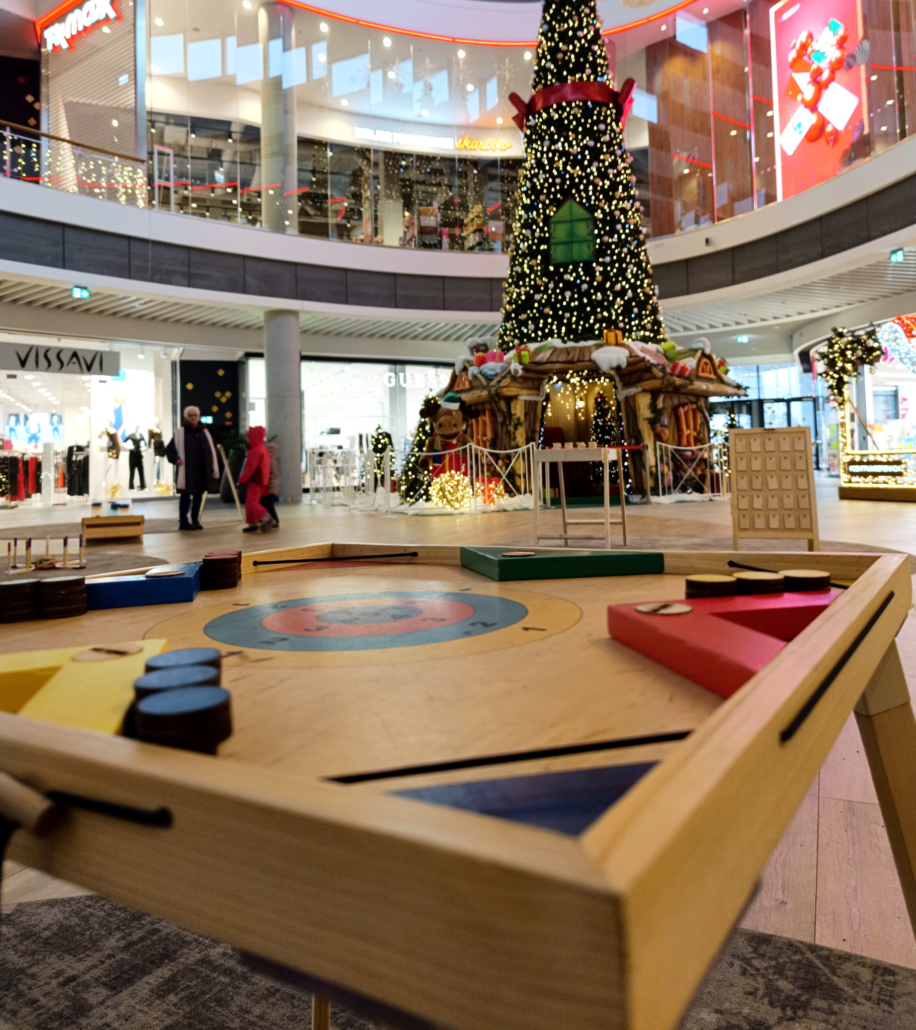 Drewniany stół do gry w kapsle z kolorowymi polami i pionkami, ustawiony w galerii handlowej z dekoracjami świątecznymi w tle.