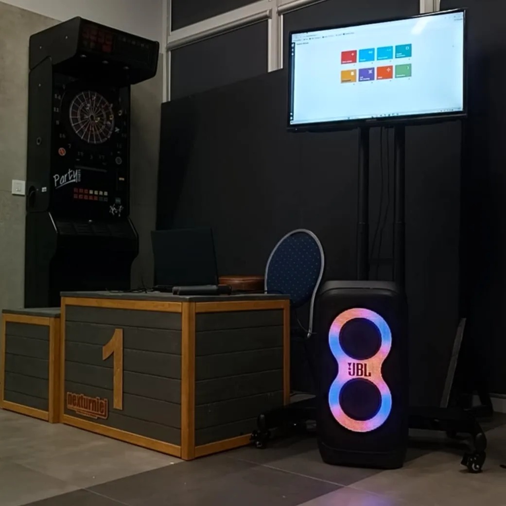Stanowisko DJ-a z drewnianą konsolą z napisem 'nexturniej', dartem, laptopem, krzesłem i głośnikiem JBL z podświetleniem LED, ustawione na tle czarnej ściany i wiszącego ekranu.