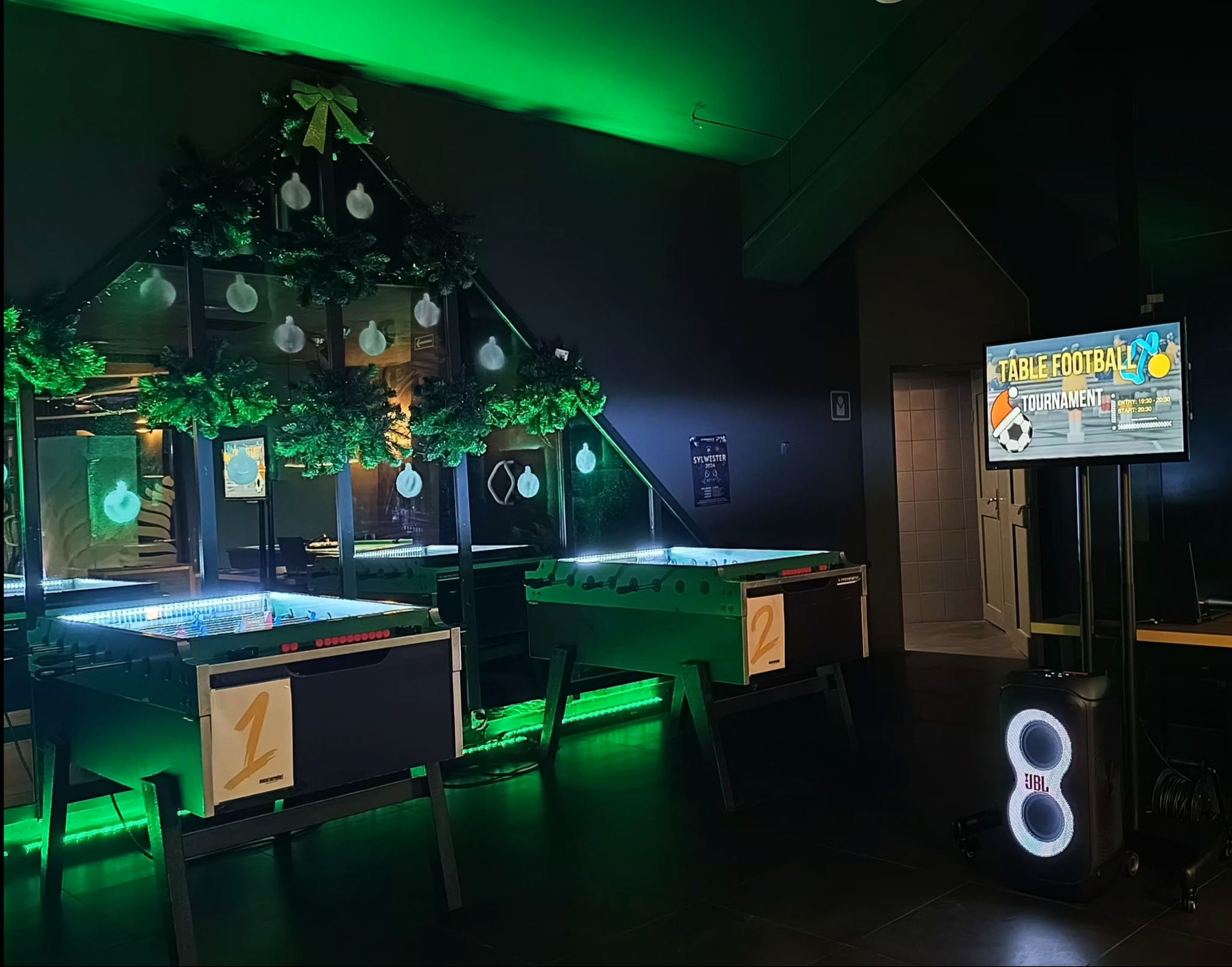 Dwa stoły do piłkarzyków z numerami 1 i 2, udekorowane zielonymi girlandami i bombkami, podświetlone zielonym światłem, obok ekran z informacją o turnieju piłkarzyków i głośnik JBL.