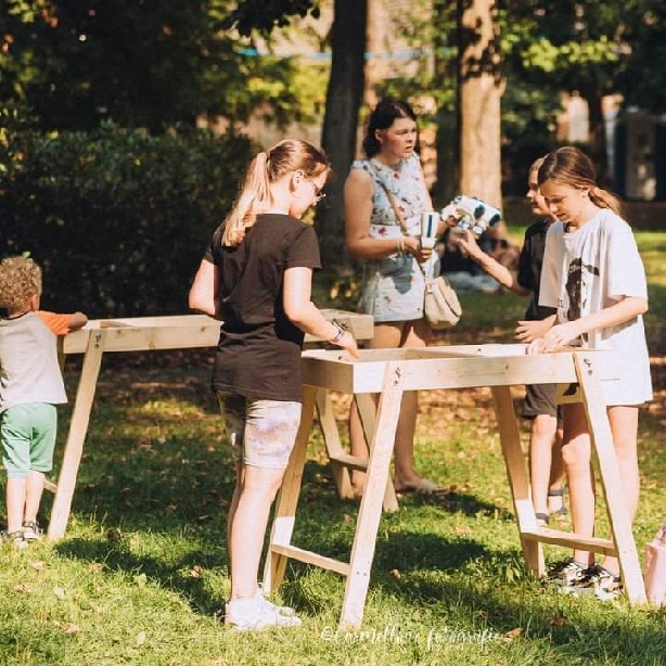 Dzieci uczestniczą w warsztatach plastycznych na świeżym powietrzu, pracując przy drewnianych stołach w parku. W tle widoczna kobieta z napojami.