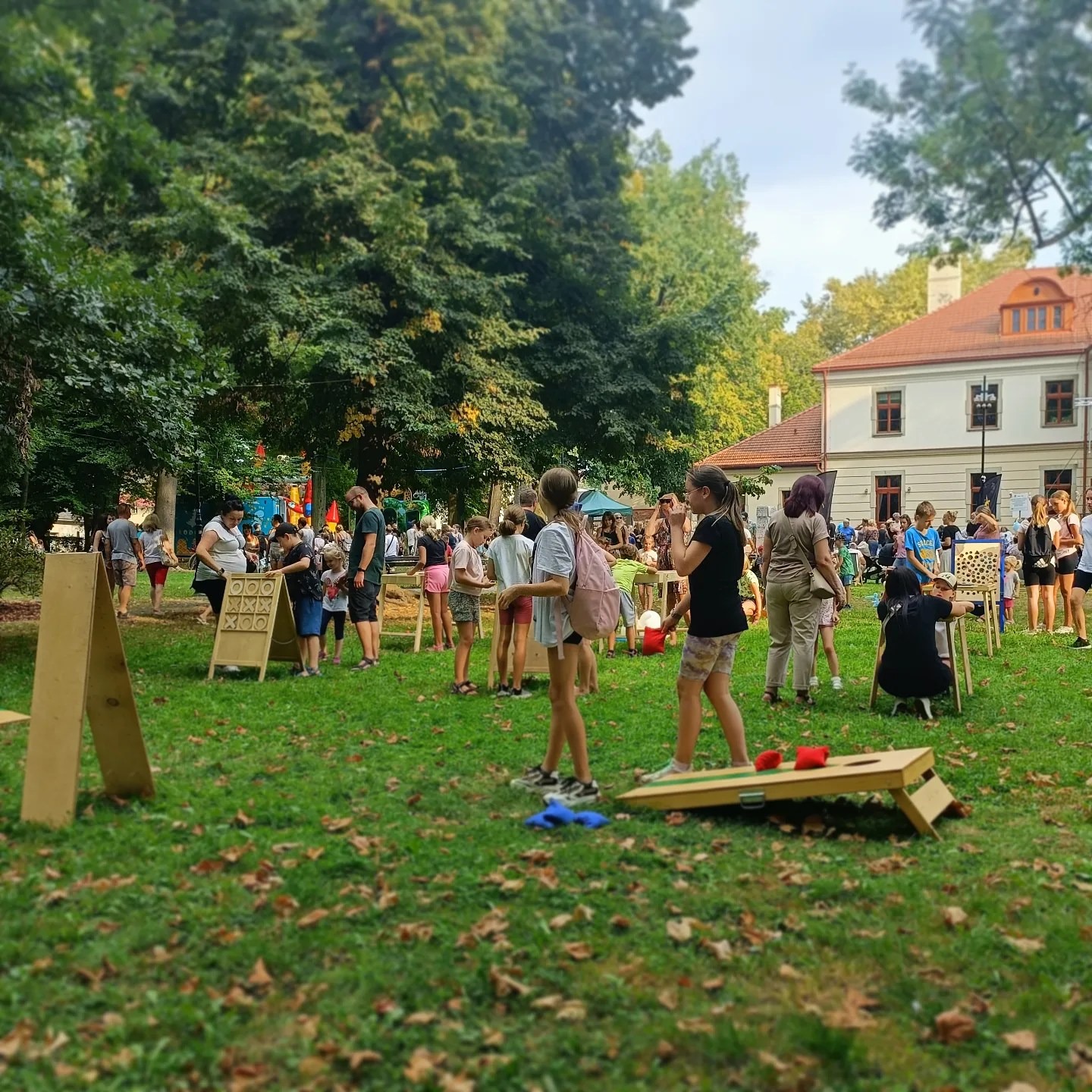 Plenerowa impreza z grami dla dzieci i dorosłych: rzut woreczkami do celu, kółko i krzyżyk, w tle budynek z czerwoną dachówką i drzewa.