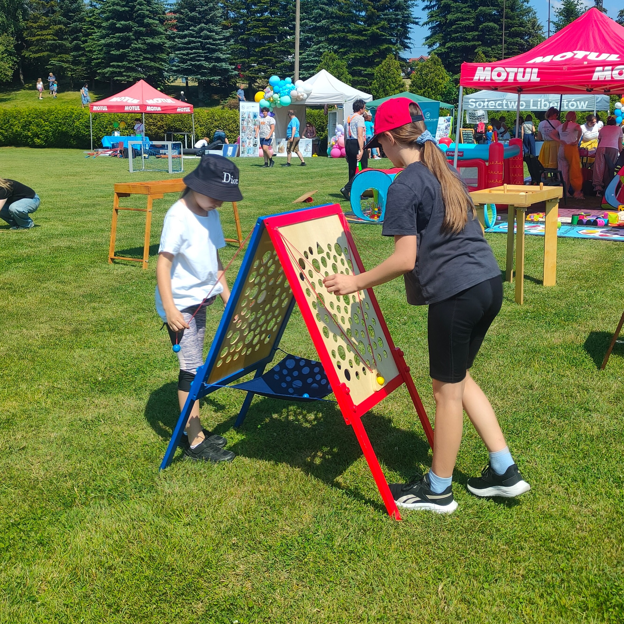 Dwoje dzieci bawi się interaktywną, drewnianą zabawką z otworami i kulkami na trawniku podczas plenerowej imprezy.