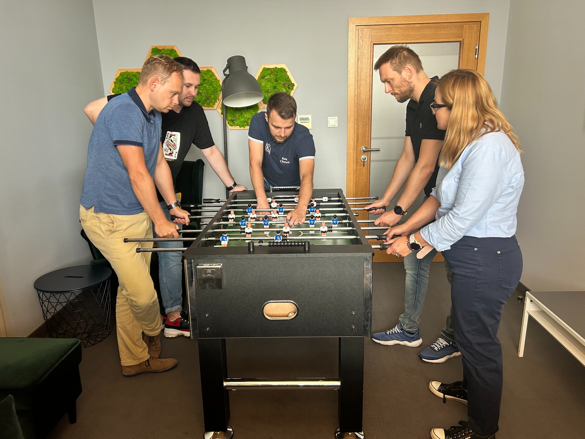 Grupa sześciu osób gra w piłkarzyki w biurze, z dekoracją z mchu w tle i lampą biurkową.