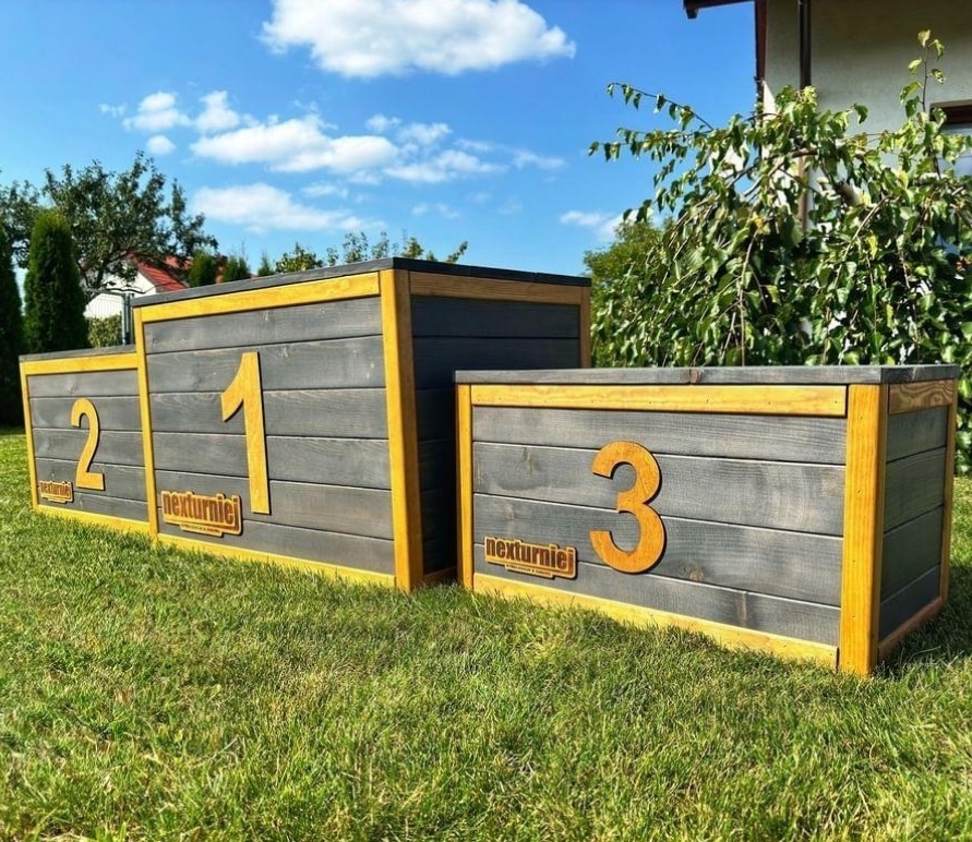 Drewniane podium z numerami 1, 2 i 3, pomalowane na szaro z żółtymi obramowaniami, stojące na trawie przed domem w słoneczny dzień.