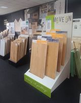 Ekspozycja paneli podłogowych w sklepie, z widocznymi różnymi odcieniami drewna i etykietami informacyjnymi na stojaku. W tle widoczne inne materiały wykończeniowe i oświetlenie sklepowe.