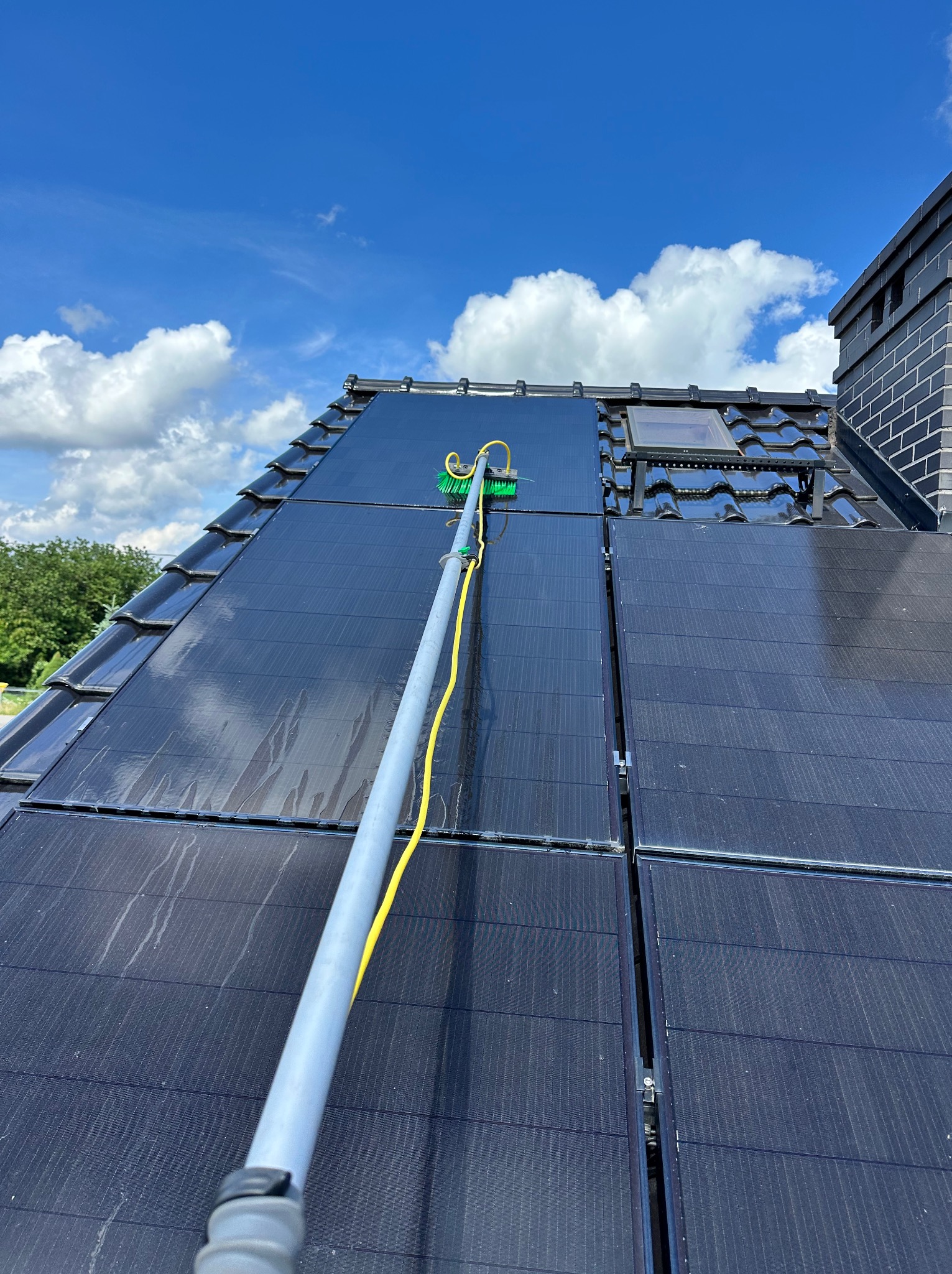 Szczotka na teleskopowym kiju czyści panele słoneczne na dachu z czarnej dachówki, błękitne niebo z chmurami w tle.